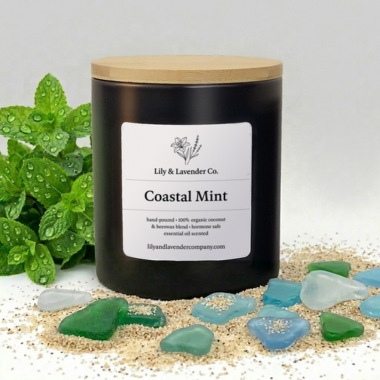 Candle labeled 'Coastal Mint' by Lily & Lavender Co. who makes Non-toxic candles made of all-natural beeswax & coconut was blend and scented with essential oils. The Candle vessel sits near some mint leaves as well as sand and sea glass.